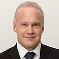 Professional portrait of a smiling man in a dark suit and tie against a light background.